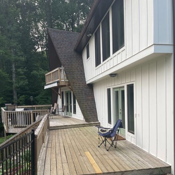 A-frame house with a wooden deck, folding chair, and forest backdrop.