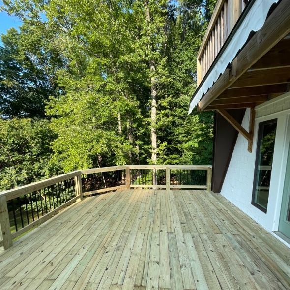 Wooden deck with railing, connected to a white house with green doors, surrounded by trees.