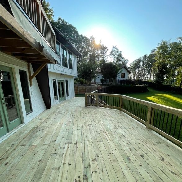 Wooden deck with railing attached to a white house with large windows. Sunny, daytime.