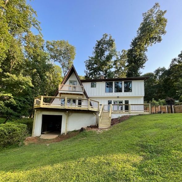 White A-frame house with a wooden deck and open carport on a grassy hill, under a blue sky.