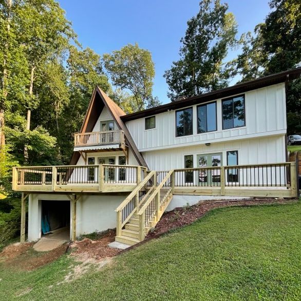 White A-frame house with a wooden deck and staircase, set in a grassy area surrounded by trees.