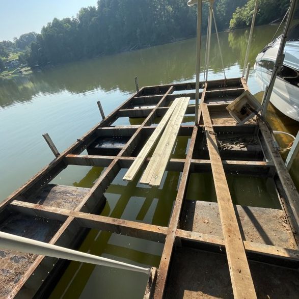 Metal dock frame on water with lumber. A boat is docked on the right. Trees line the background.