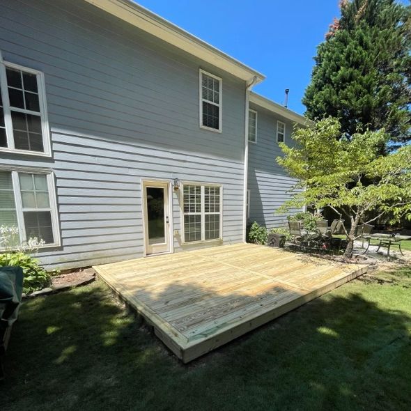 A newly constructed wooden deck extends from the back of a light blue house, surrounded by green grass and a small tree.