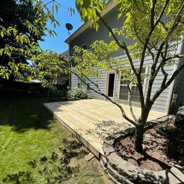 Backyard with a wooden deck, a small tree, and a house with a sliding glass door. Sunny day.