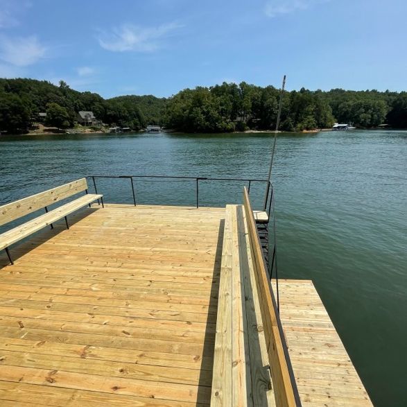 Wooden dock with benches and railings, overlooking calm water and wooded shoreline on a sunny day.