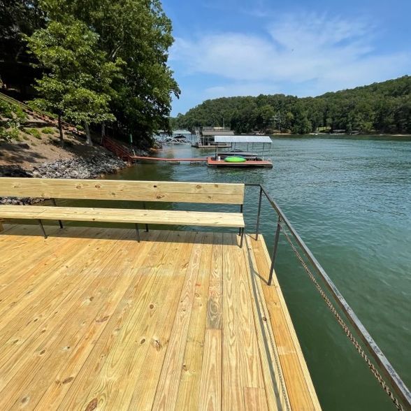 Wooden dock with bench overlooking a lake, boat docks, and lush green trees. Sunny day.