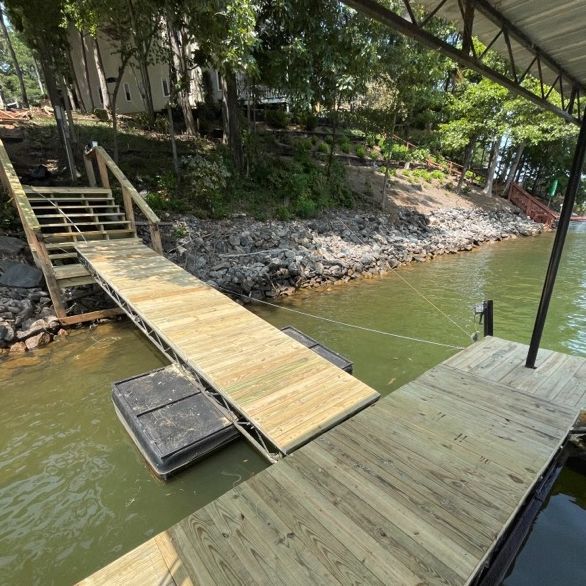 Wooden dock and ramp leading to lake. Green water, trees, and a house in the background.