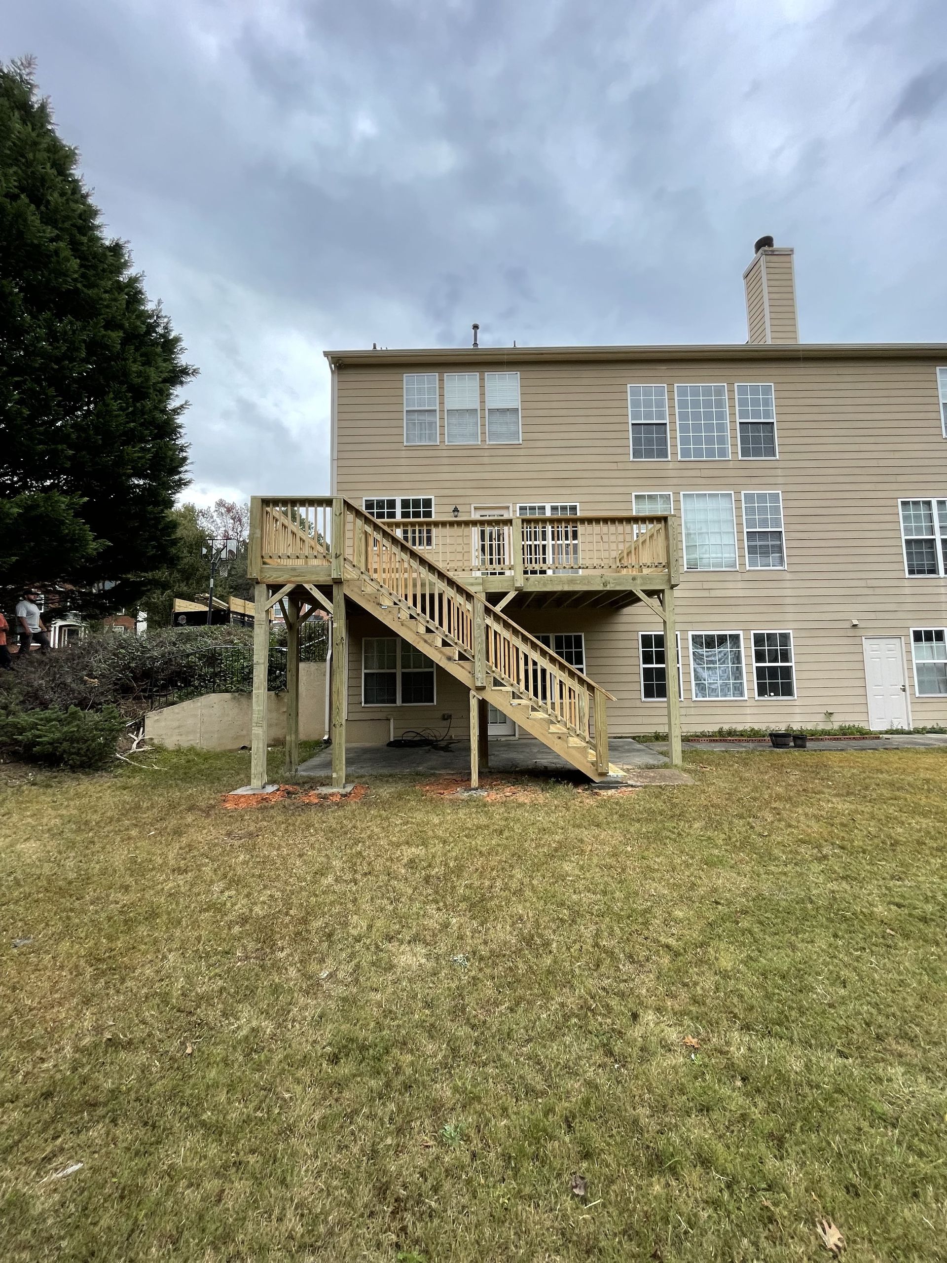 Wooden deck with stairs attached to a beige two-story house, set in a grassy yard under a cloudy sky.