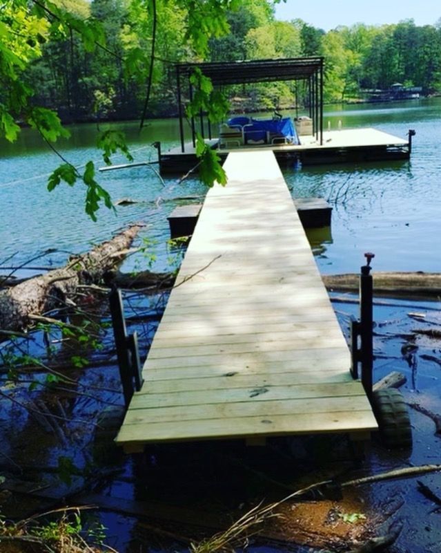 Wooden dock extending into a lake, with a canopy-covered platform at the end. Green trees and foliage surround the water.