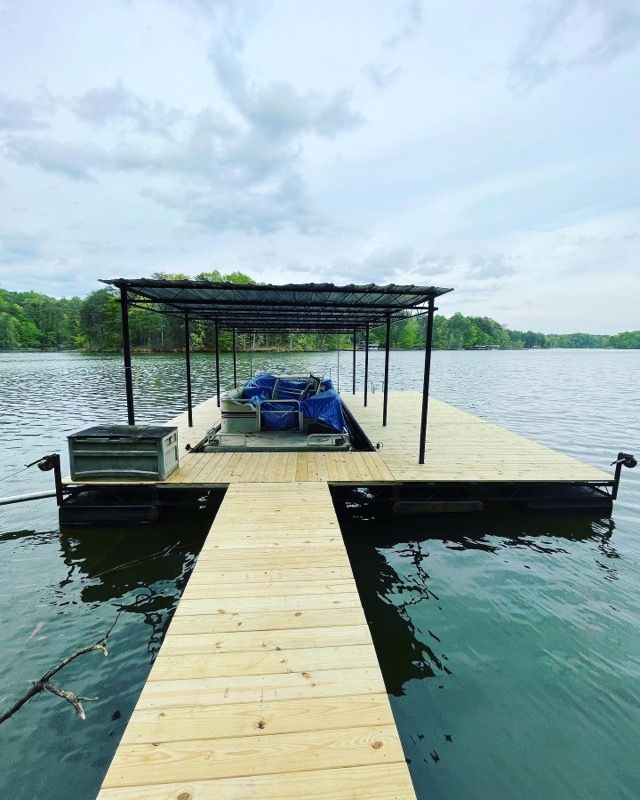 Wooden dock on a lake with a boat covered by a canopy. Green trees line the distant shore, cloudy sky.