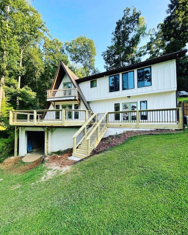 White A-frame house with new wooden deck and stairs, set in a green, grassy yard.