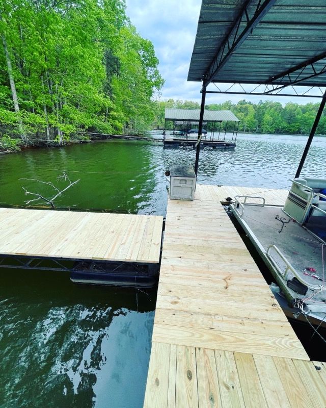 Wooden dock on a lake, boat tied alongside. Green trees and overcast sky in the background.