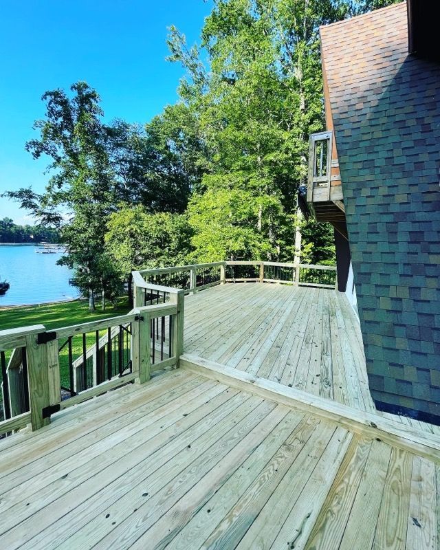 Wooden deck overlooking a lake and trees, attached to a house with a brown roof.