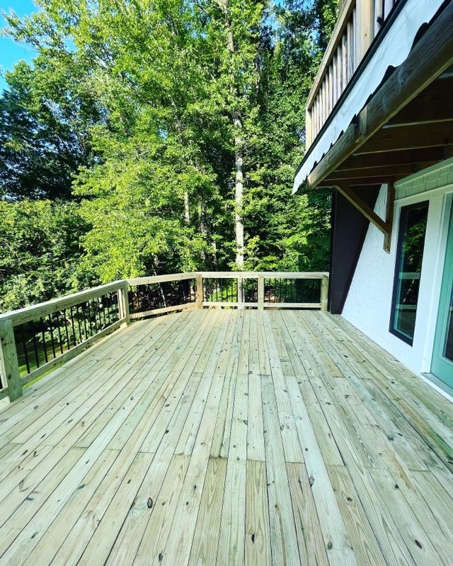 Wooden deck with a railing, next to a building with a balcony, trees in the background.
