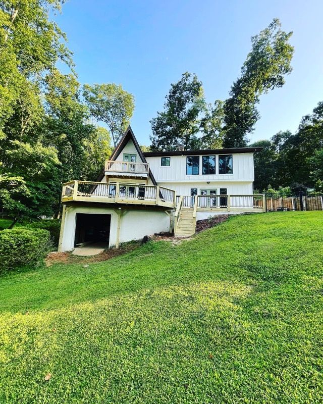 White A-frame house on a grassy hill, with a deck and garage. Trees frame the house against a blue sky.