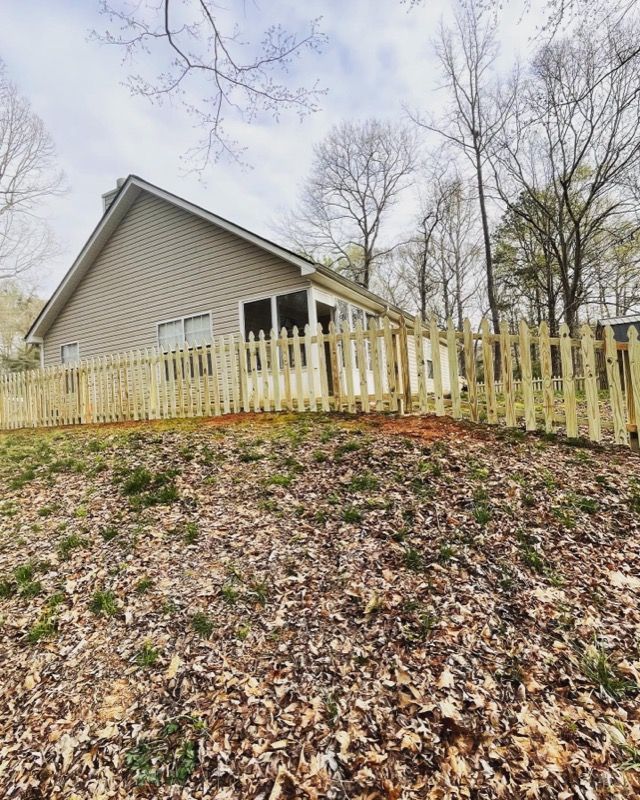 Wooden picket fence surrounding a light beige house on a grassy hill; overcast sky.