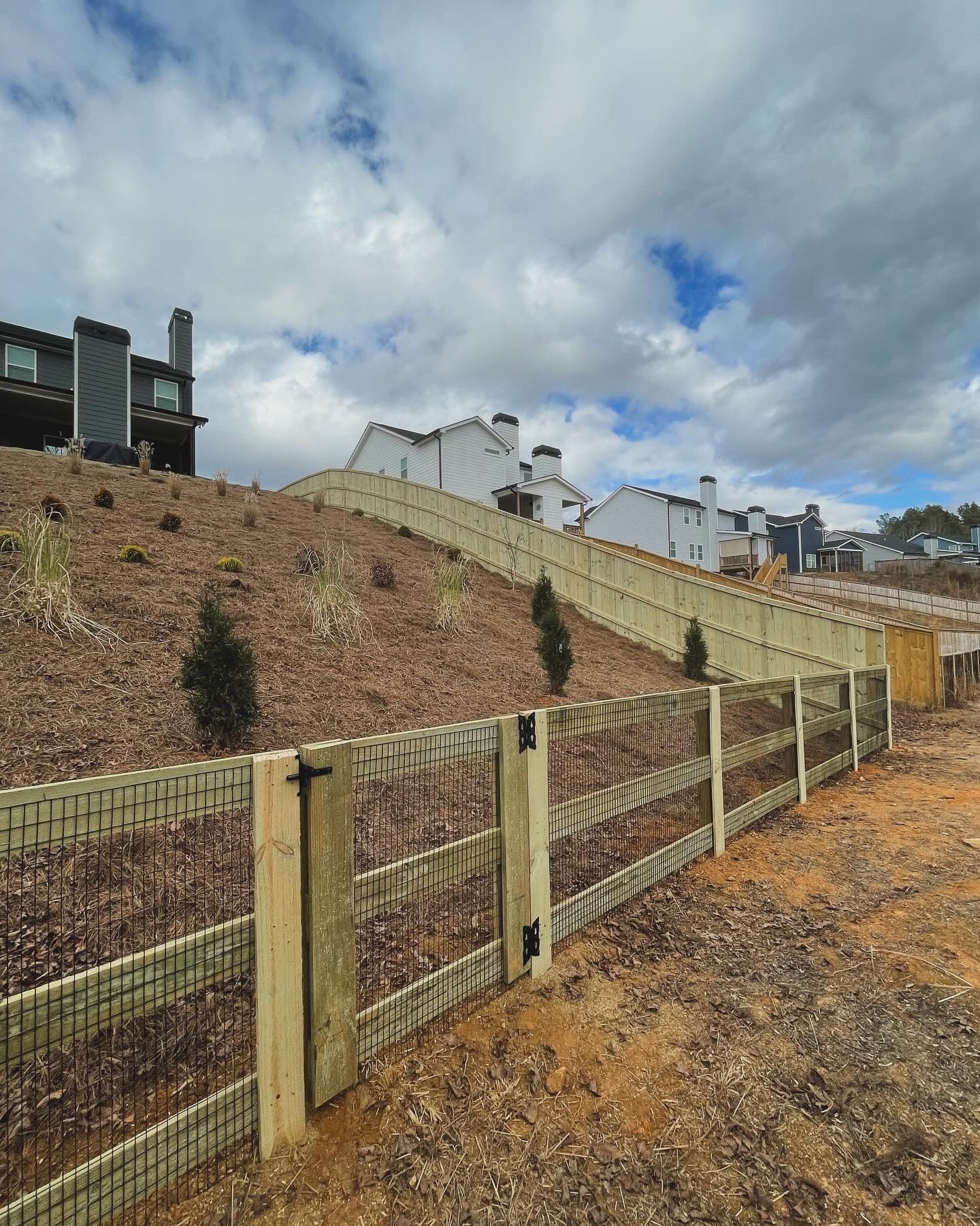 A sloped hillside with a new wooden fence, houses in the background, under a cloudy sky.