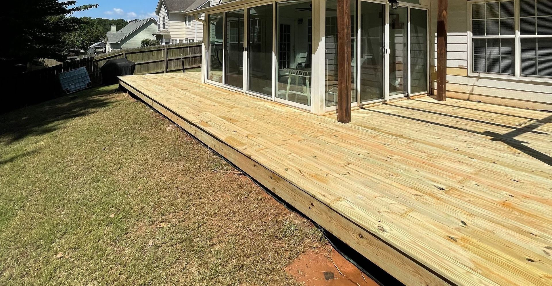 Wooden deck extending from a house with glass sliding doors, built on a grassy lawn under a sunny sky.