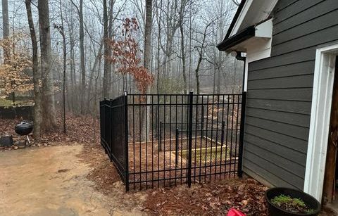Black metal fence enclosing a small yard, next to a gray building. Bare trees in background.
