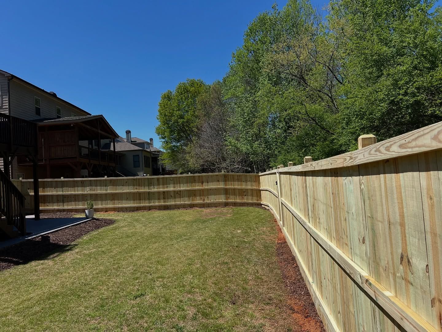 Backyard with patchy grass, wooden fence, and trees under a bright blue sky