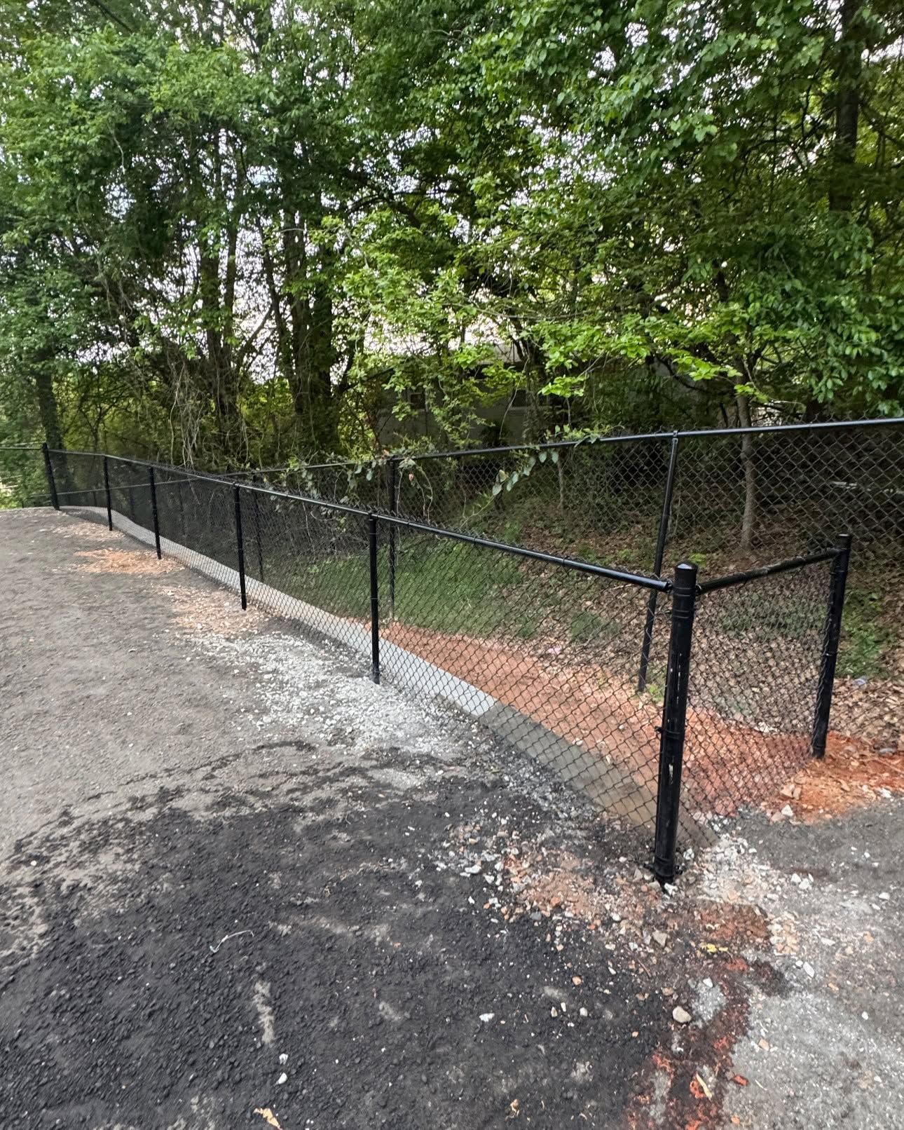 Black chain-link fence along a gravel path beside dense green trees and shrubs.