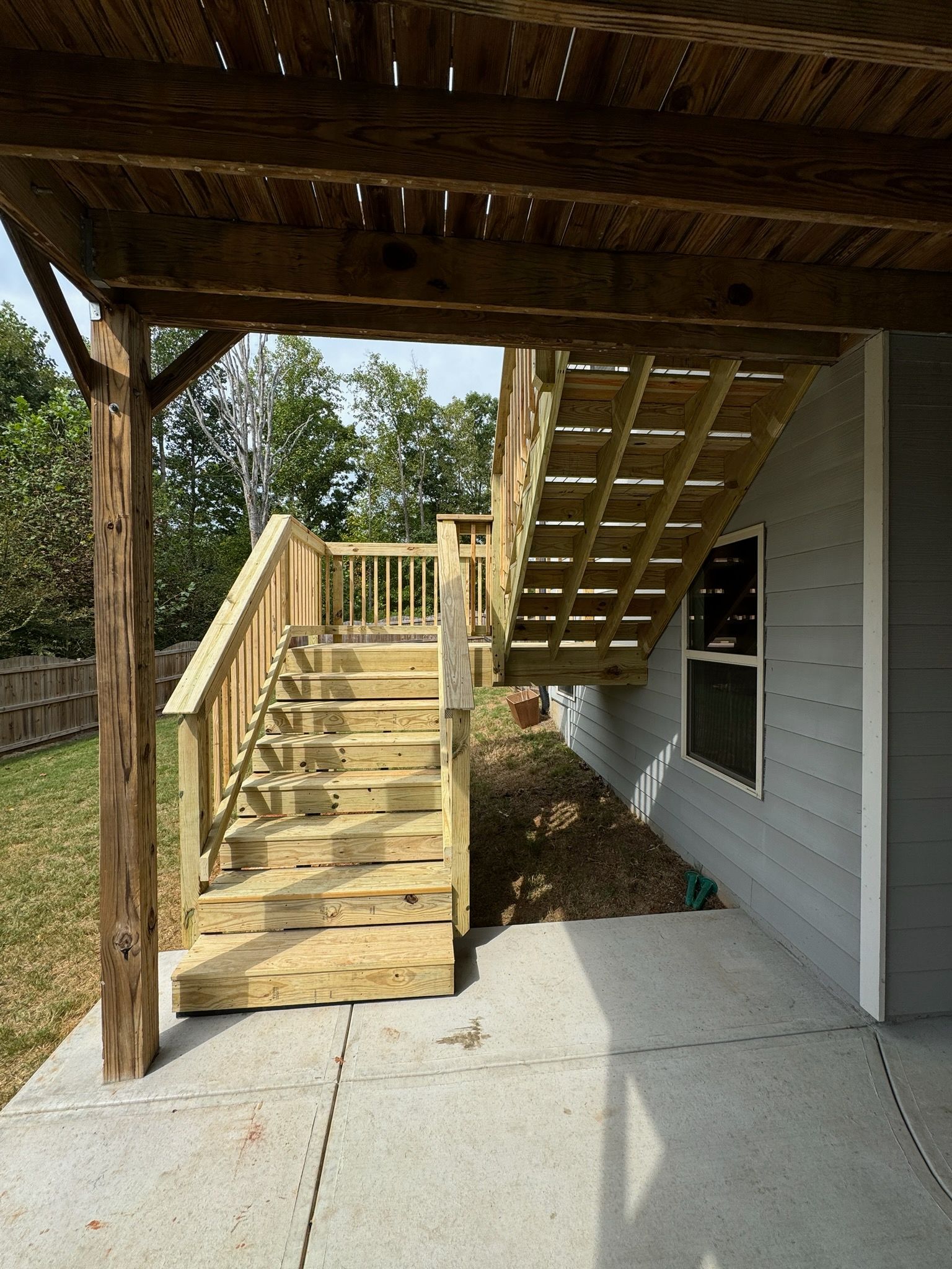 Wooden staircase leading up to a deck under a pergola next to a house with a window.