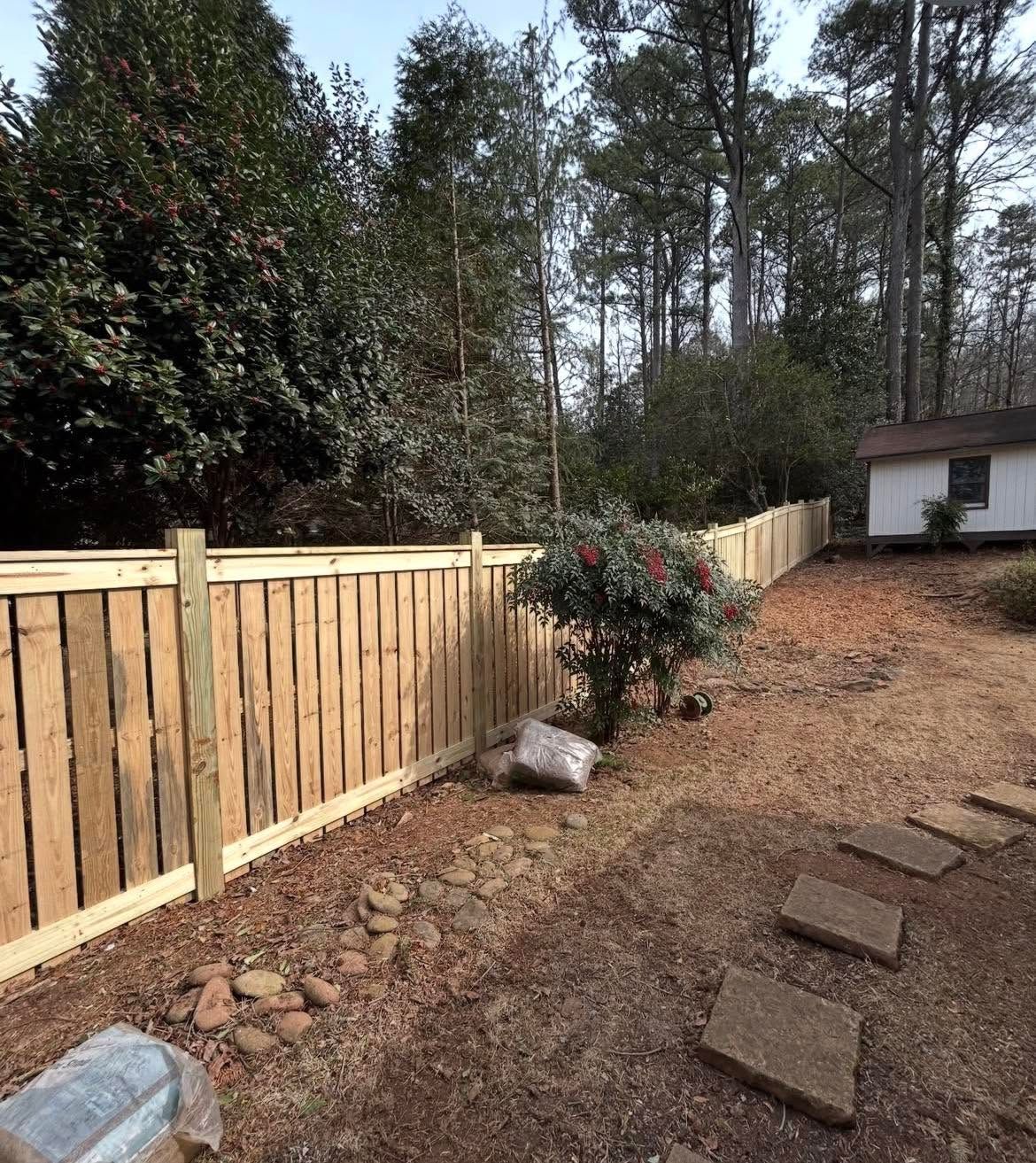 Wooden privacy fence along a gravel yard with stepping stones, shrubs, and tall pine trees in the background