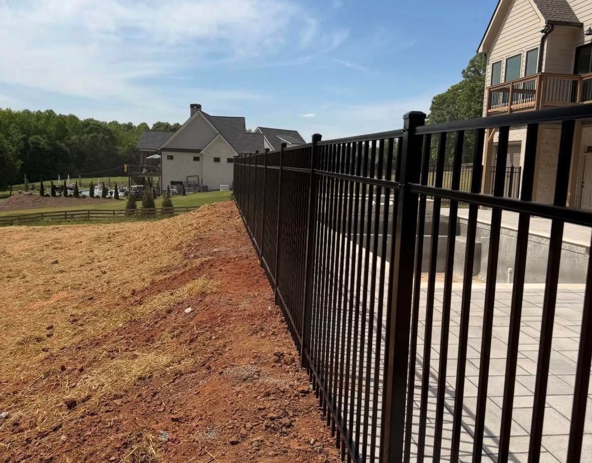 Black metal fence beside a paved driveway, with a house and open grassy yard on a sunny day.