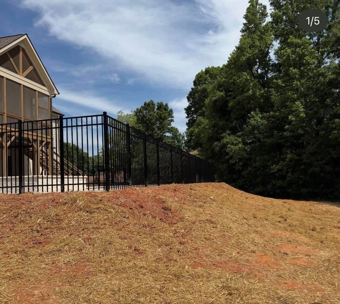 Black metal fence beside a beige house on a grassy slope, with trees and a cloudy sky.