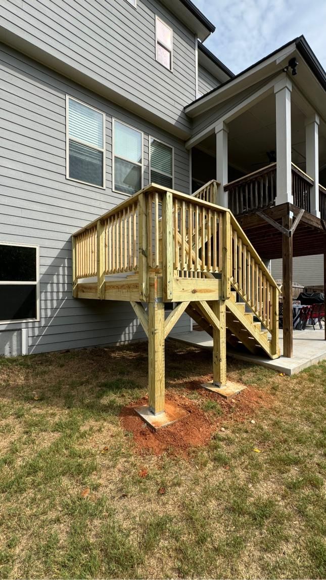 New wooden deck with stairs attached to a gray house, set on concrete footings on brown grass.