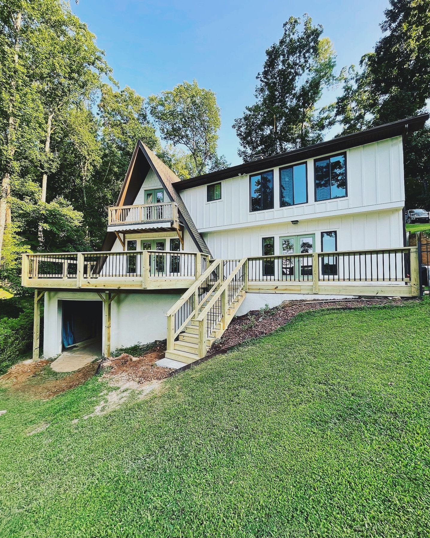 White A-frame house with large deck, surrounded by green grass and trees, under a blue sky.