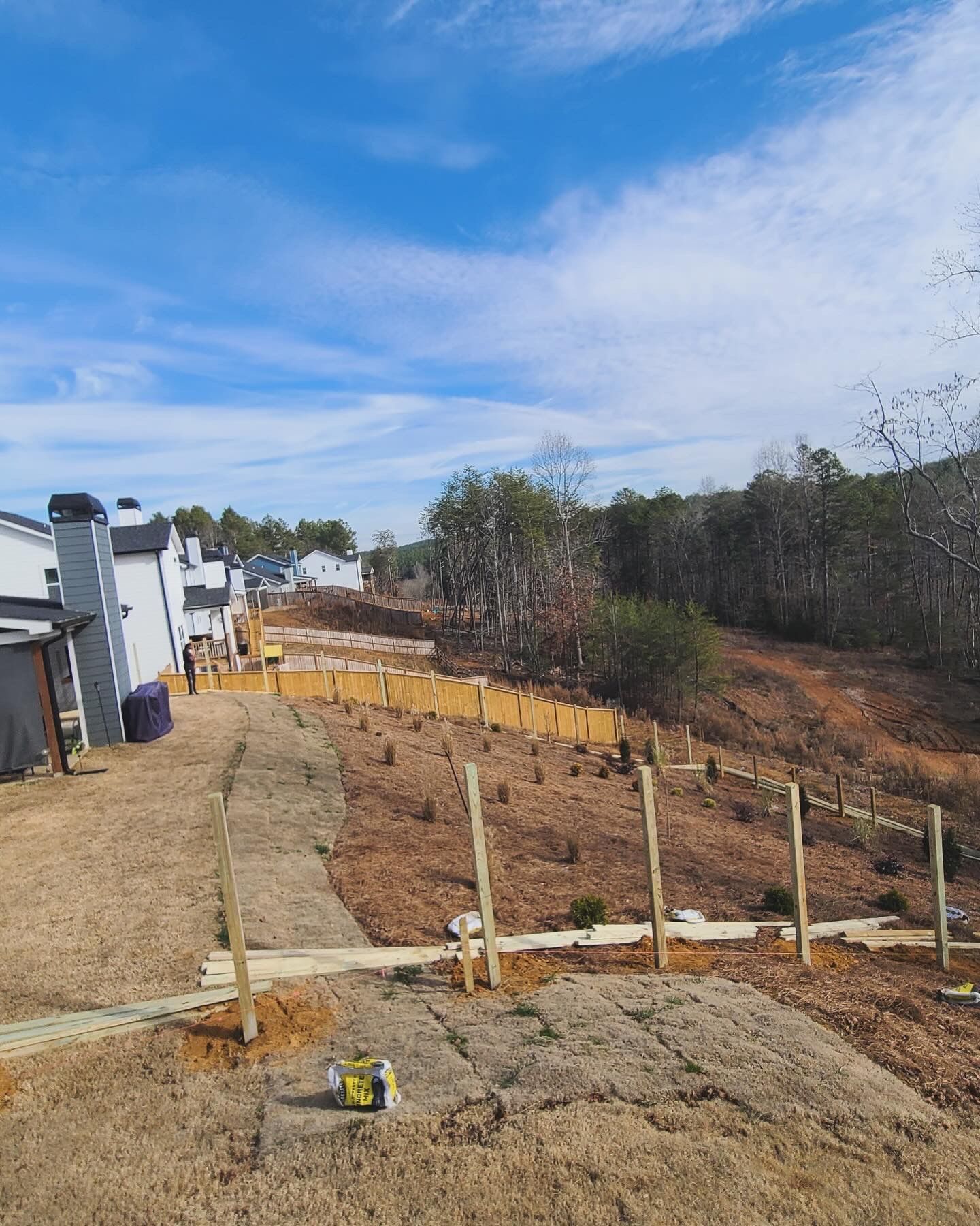 A partially constructed wooden fence lines a grassy slope on a sunny day with houses in the background.