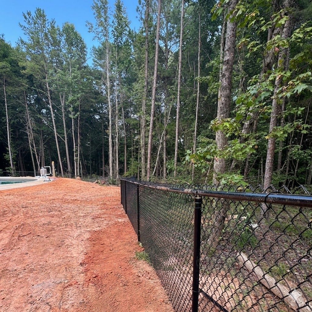Black chain-link fence beside a red dirt path and forest of tall pine trees under a blue sky