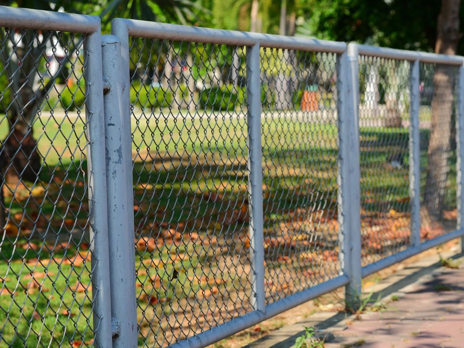 Chain-link fence beside a sidewalk in a park, with trees and grass in the background