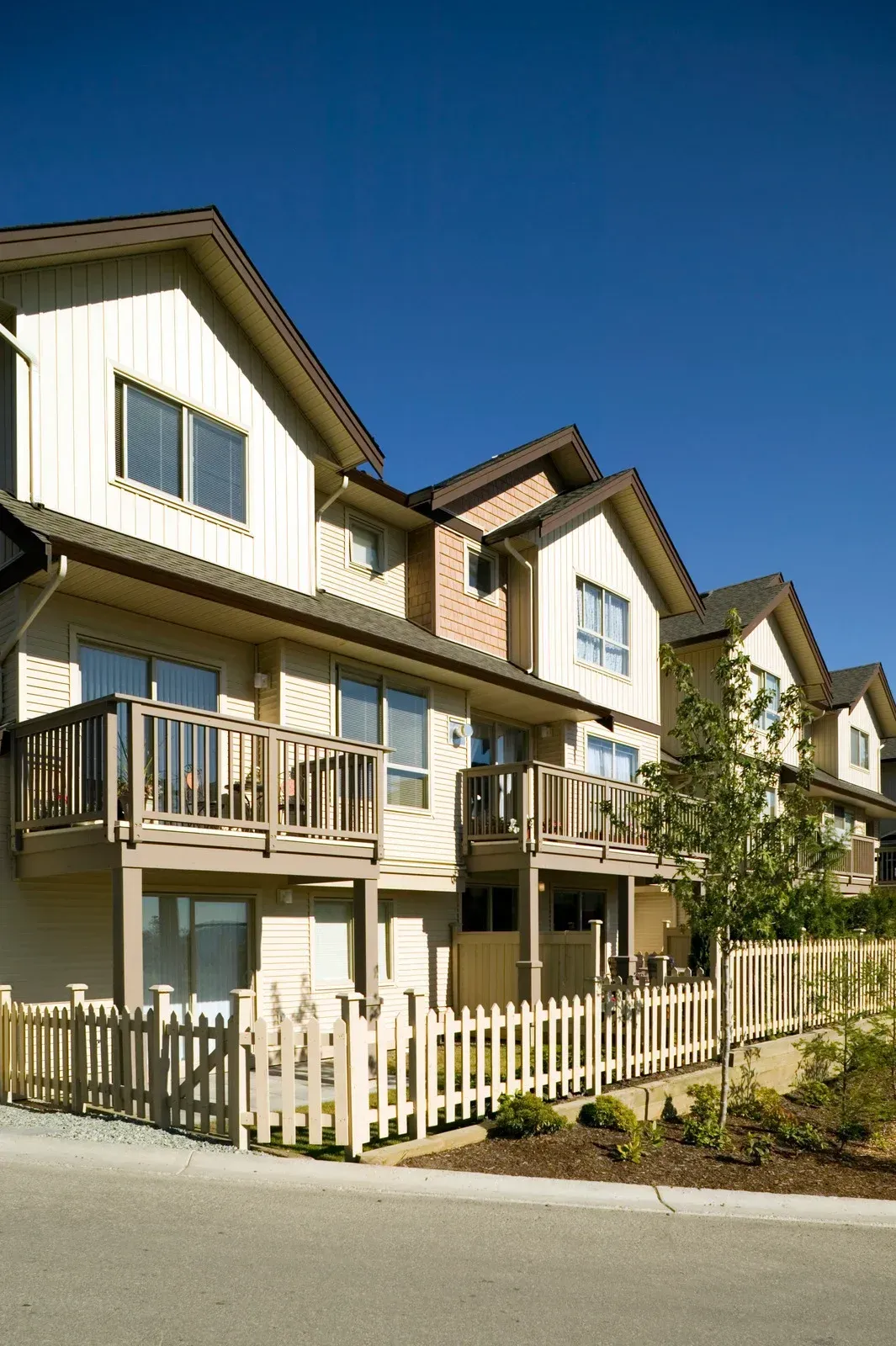 Row of beige townhouses with balconies, picket fence, and clear blue sky.