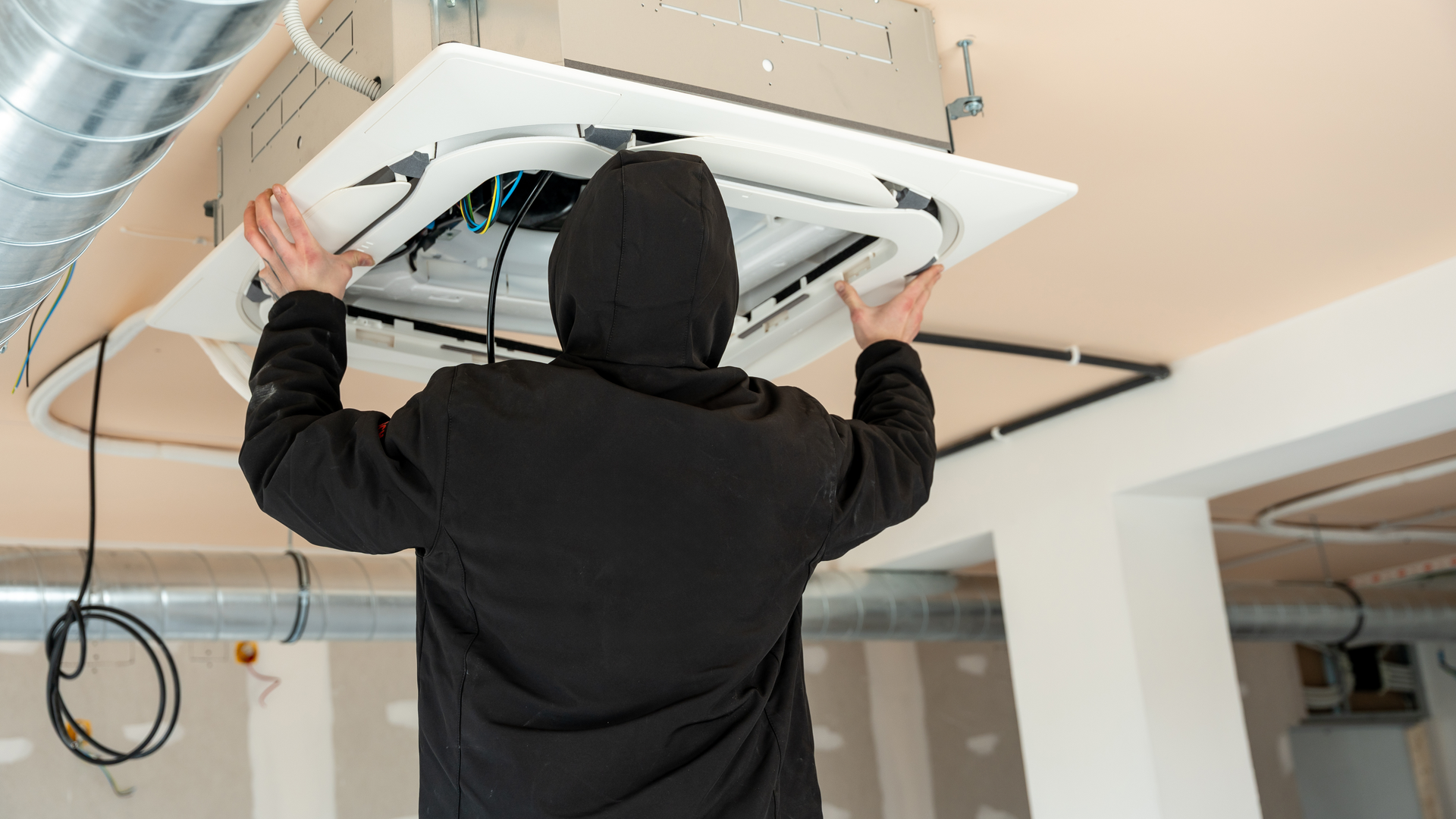 Person installing a ceiling-mounted unit with exposed wiring in a room, viewed from behind