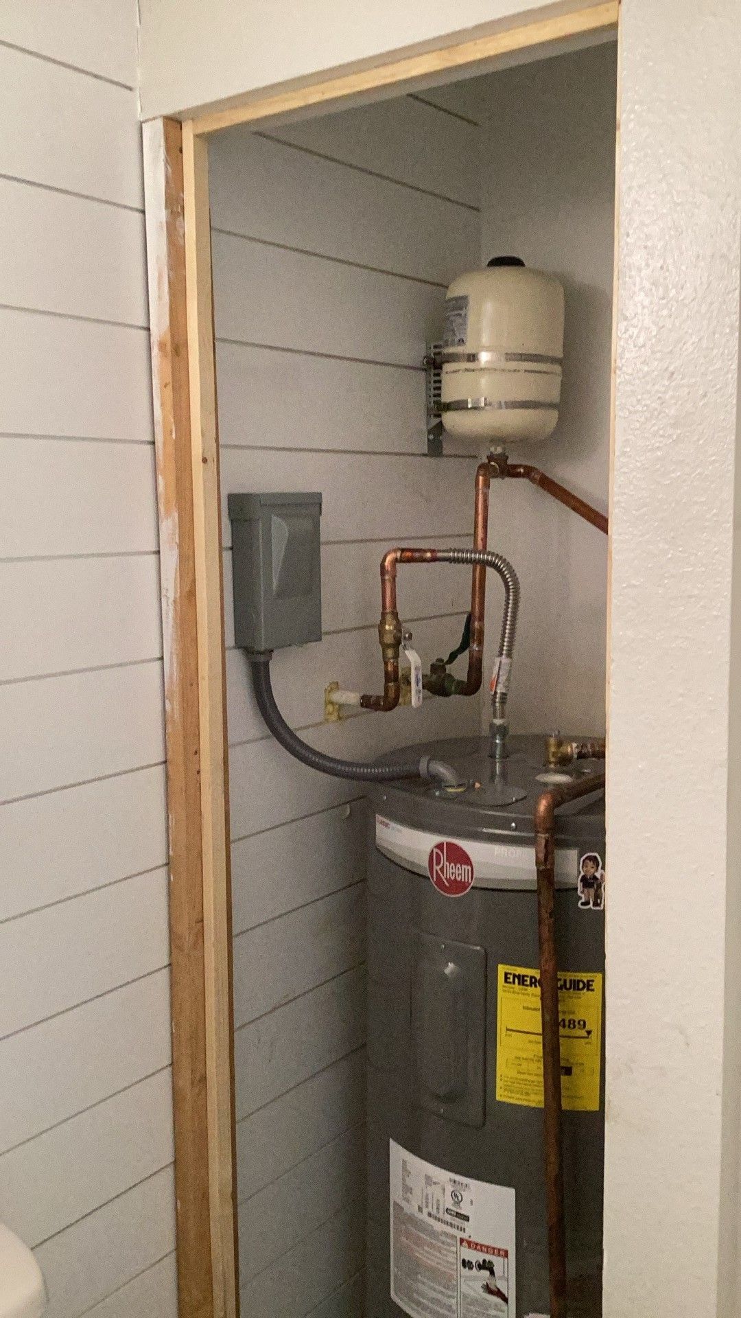 Water heater in a narrow utility closet with copper pipes and a pressure tank.