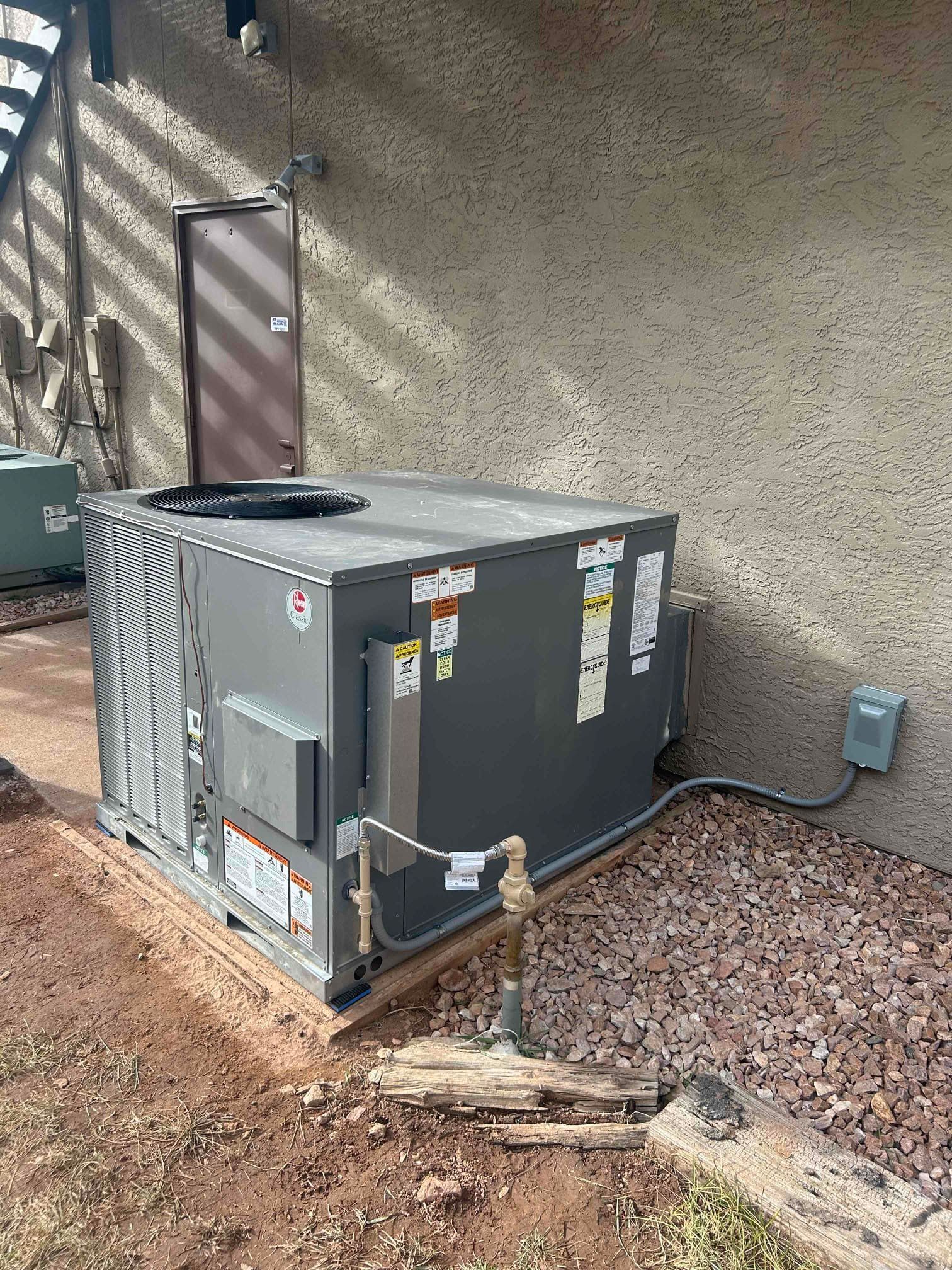 Outdoor HVAC unit beside a tan stucco wall with gravel landscaping and utility lines