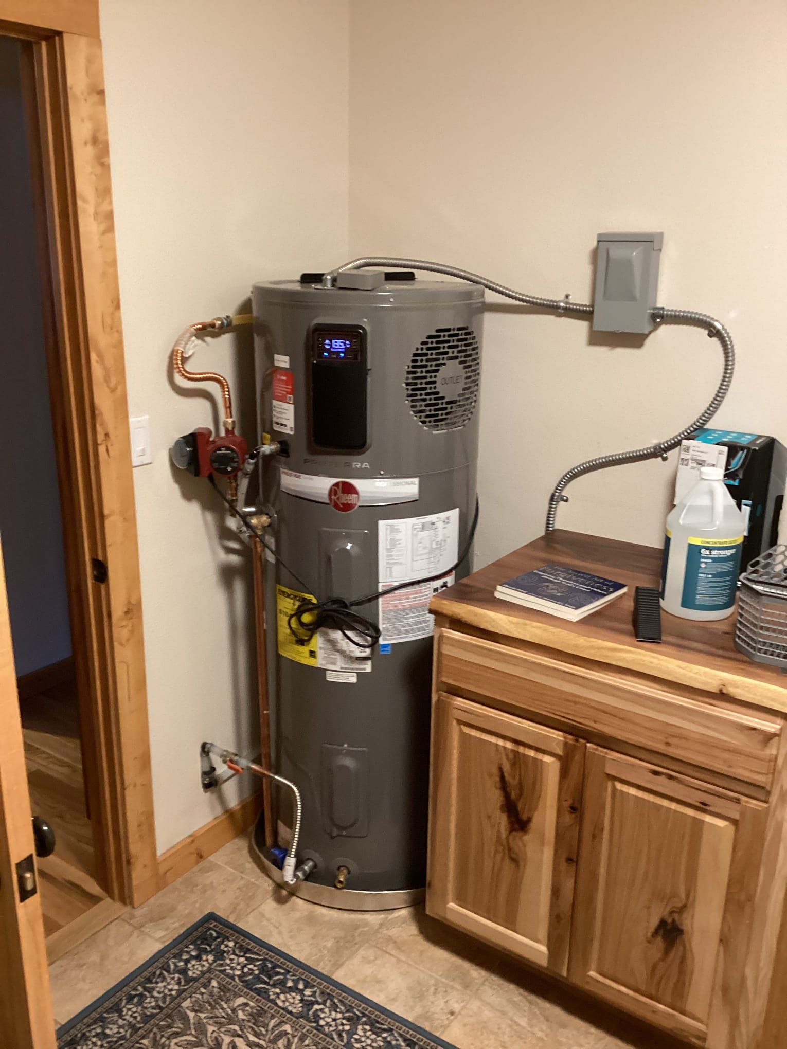 Water heater in a utility room beside a wooden cabinet and doorway, with exposed pipes and wall-mounted electrical box.