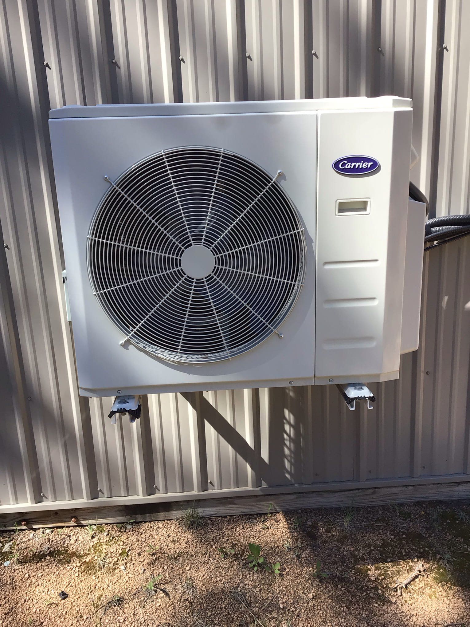 White outdoor heat pump unit mounted on a corrugated metal wall above gravel.
