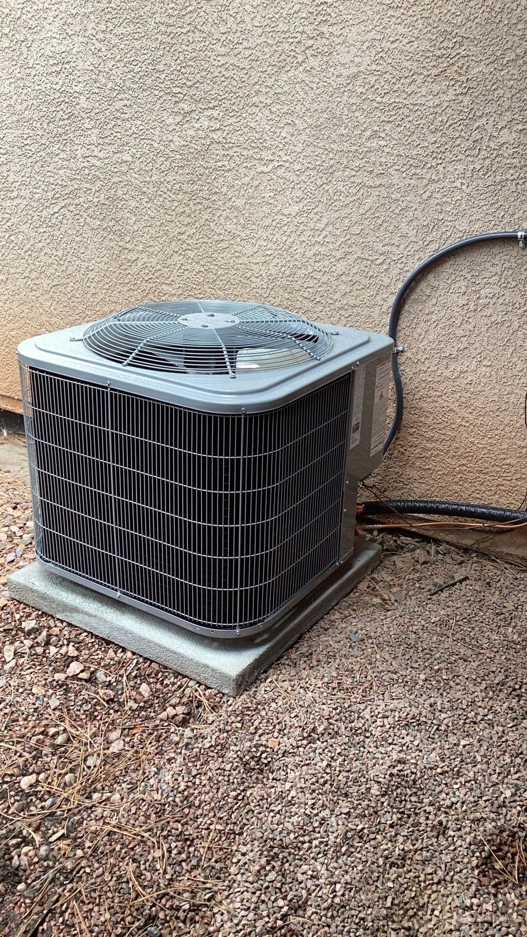 Outdoor HVAC condenser unit beside a stucco wall on gravel, with a hose or cable behind it