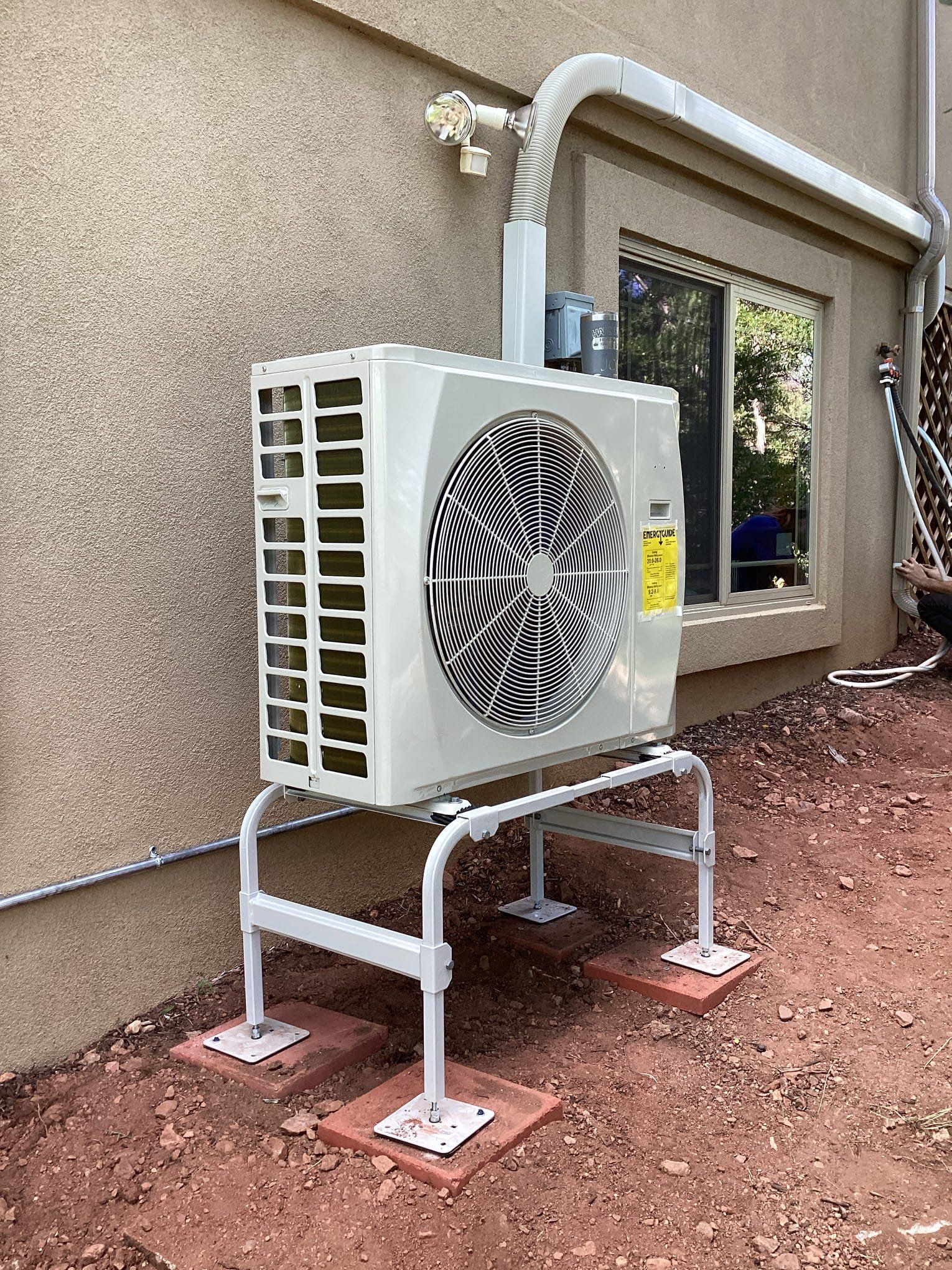 Outdoor HVAC condenser unit on a metal stand beside a stucco house wall, with pipes and a window nearby.