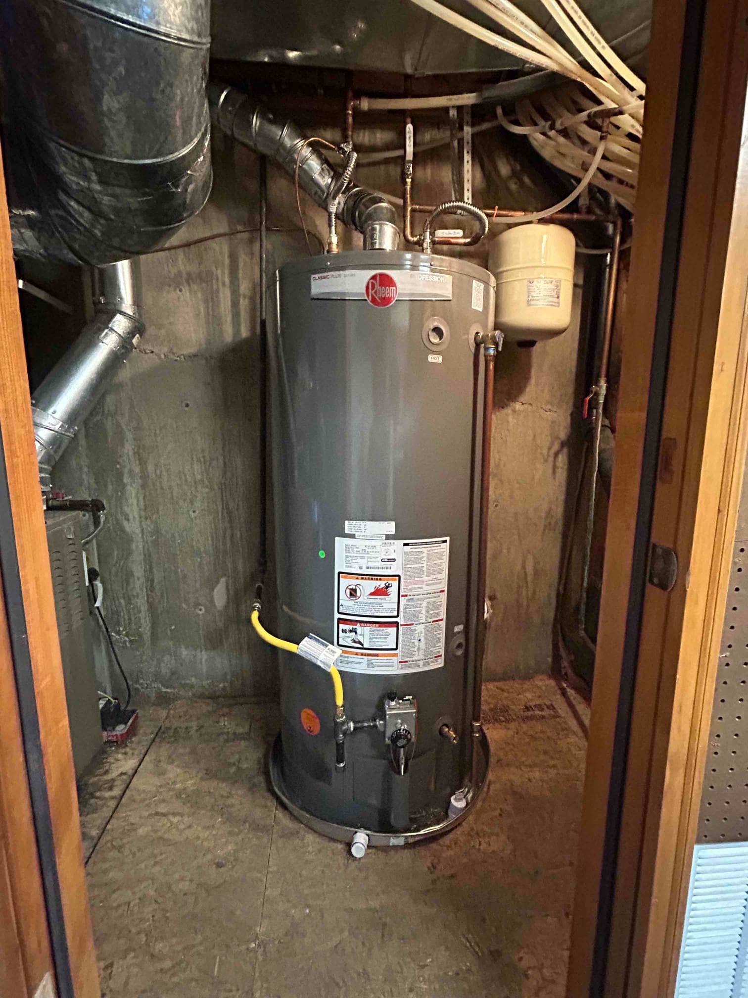 Water heater in a cramped utility closet with exposed pipes and wiring.