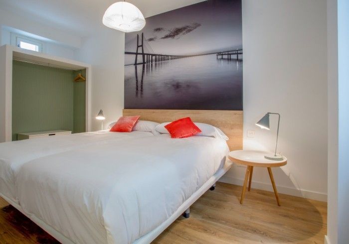 Dormitorio minimalista con cama blanca, almohadas rojas, lámpara de noche y cuadro con forma de puente.