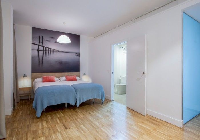 Habitación luminosa con una colcha azul, suelos de madera, cuadros en las paredes y una puerta abierta que da al baño.