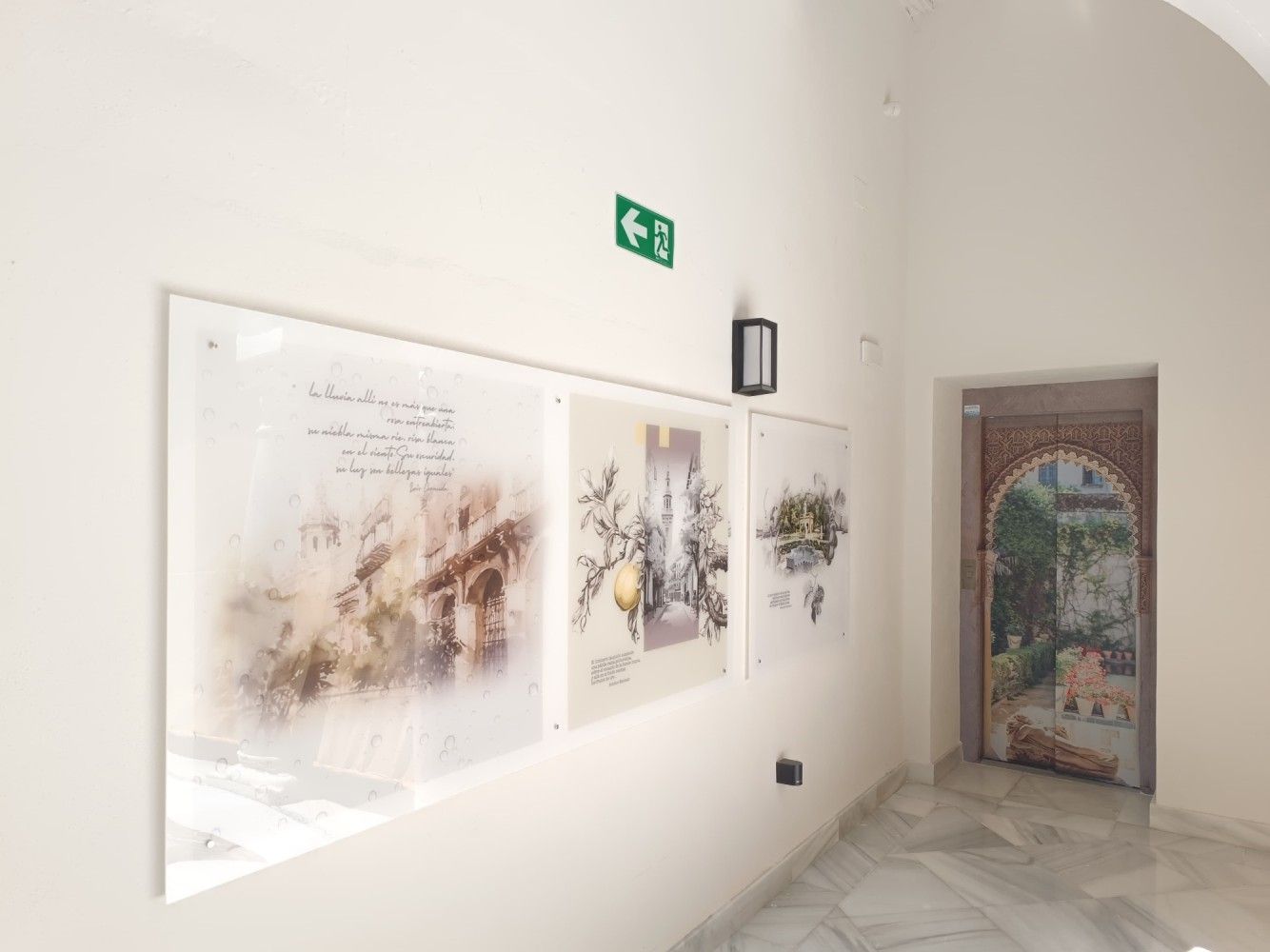 Pasillo blanco de la galería con arte abstracto enmarcado, un letrero de salida verde y una puerta arqueada ornamentada.