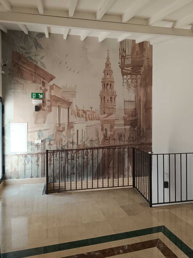 Pasillo con mural en sepia de una calle y torre históricas, barandilla negra y suelo de baldosas.