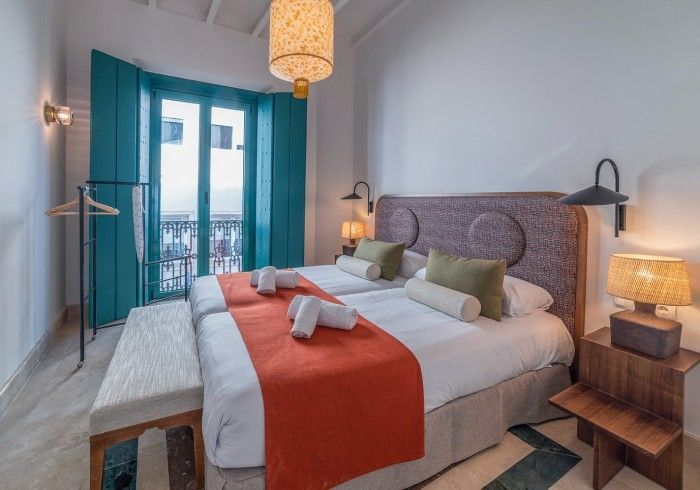Dormitorio moderno con cortinas color turquesa, cama gris, manta naranja y lámparas de noche.