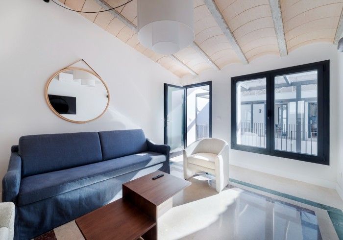 Sala de estar moderna con sofá azul, silla blanca, espejo, mesa de centro y grandes ventanales abiertos.
