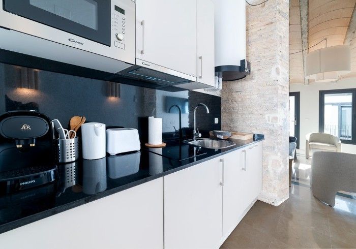 Cocina moderna con gabinetes blancos, encimeras negras, electrodomésticos y una puerta luminosa que da a la sala de estar.